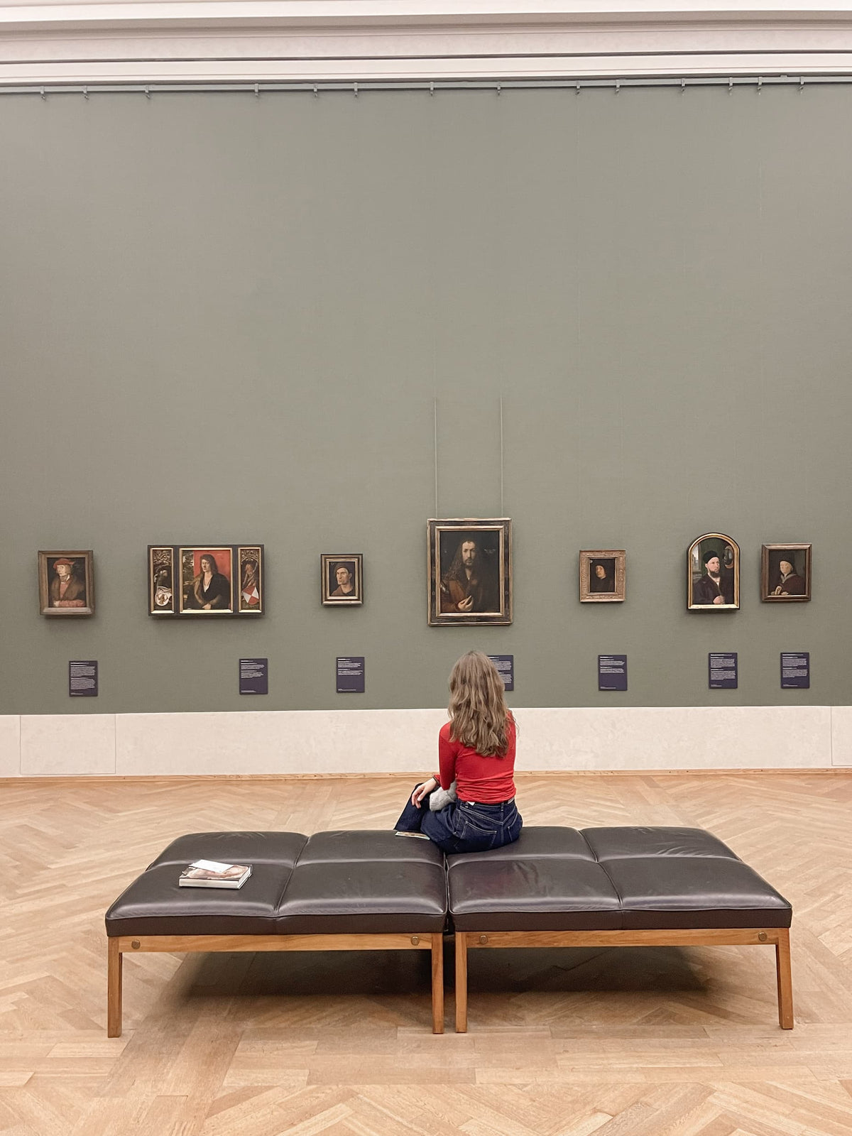 A woman in a red top sits alone on a bench in a museum gallery, facing a wall hung with Renaissance portraits.