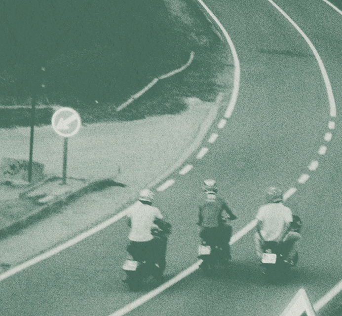 Two people riding motor scooters along a curved coastal road by the sea, seen from above
