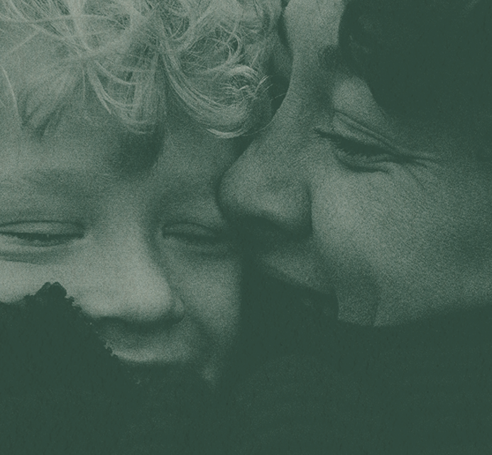 A close-up of a parent kissing a curly-haired toddler on the cheek, both smiling in a tender moment