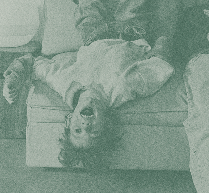 A young child lying upside down on a couch, smiling playfully at the camera with their hair hanging down