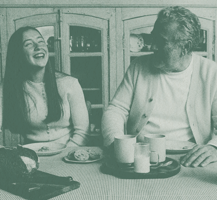 Two women laughing together at a kitchen table over coffee cups and snacks, sharing a joyful conversation