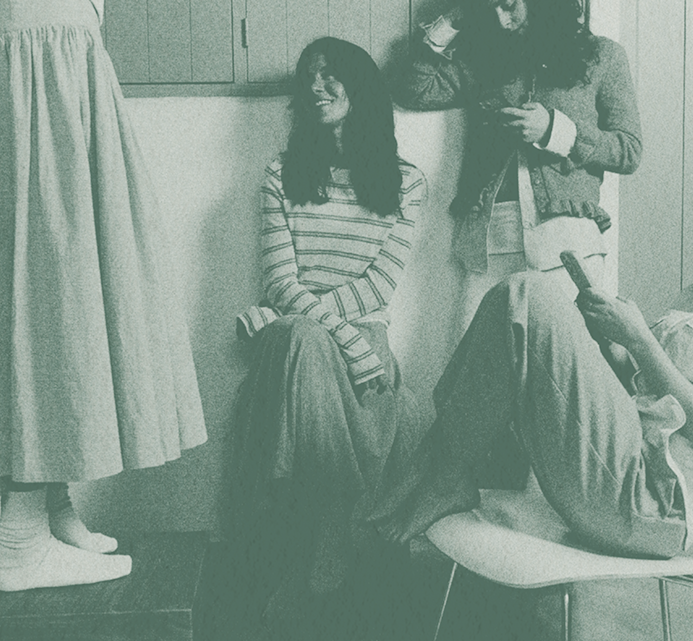 Two young women in a small kitchen or laundry room, one sitting on the counter while the other stands nearby, in a casual everyday moment