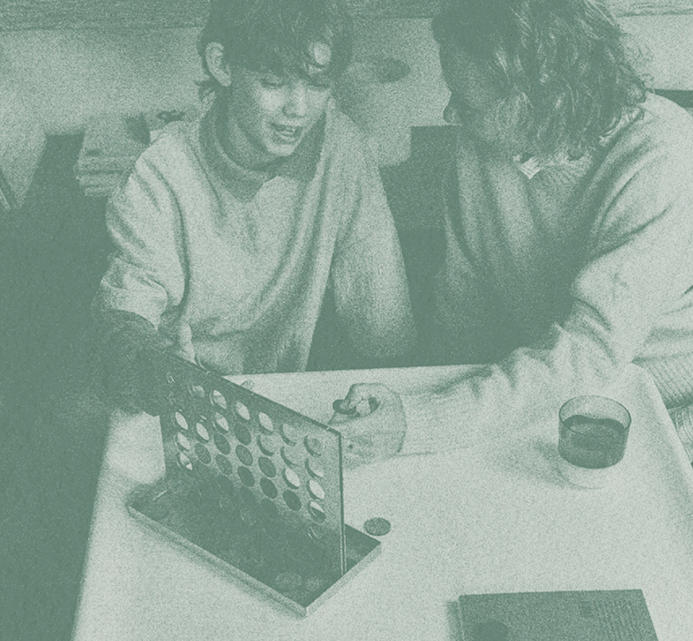 Two young people sitting at a table, leaning in together to look at a Connect Four board game, engaged in friendly competition
