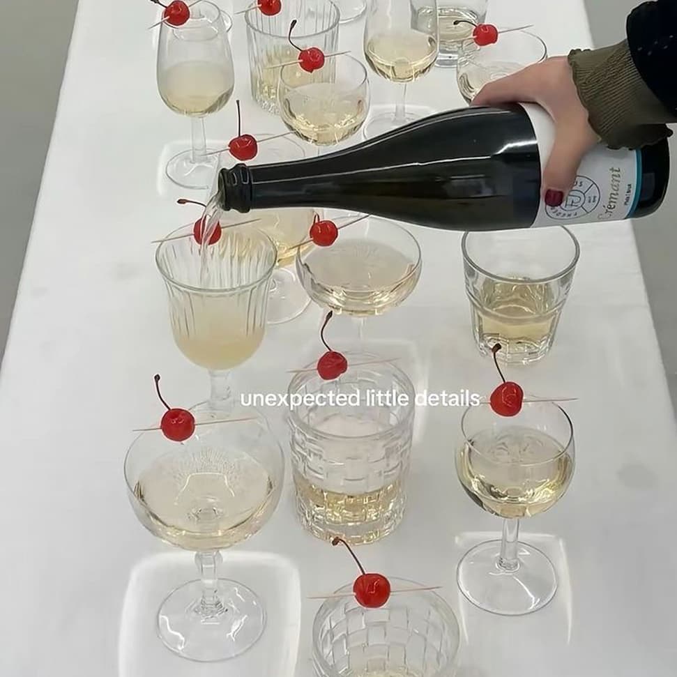 A hand pours sparkling wine into a row of mismatched glasses on a white table, each glass topped with a cocktail cherry on a stick, under the caption "unexpected little details".