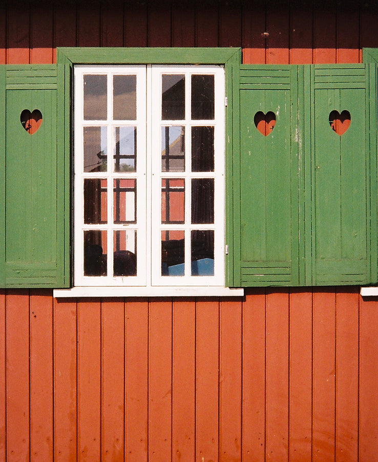 A white-framed window with green shutters decorated with heart-shaped cutouts on a red wooden building.