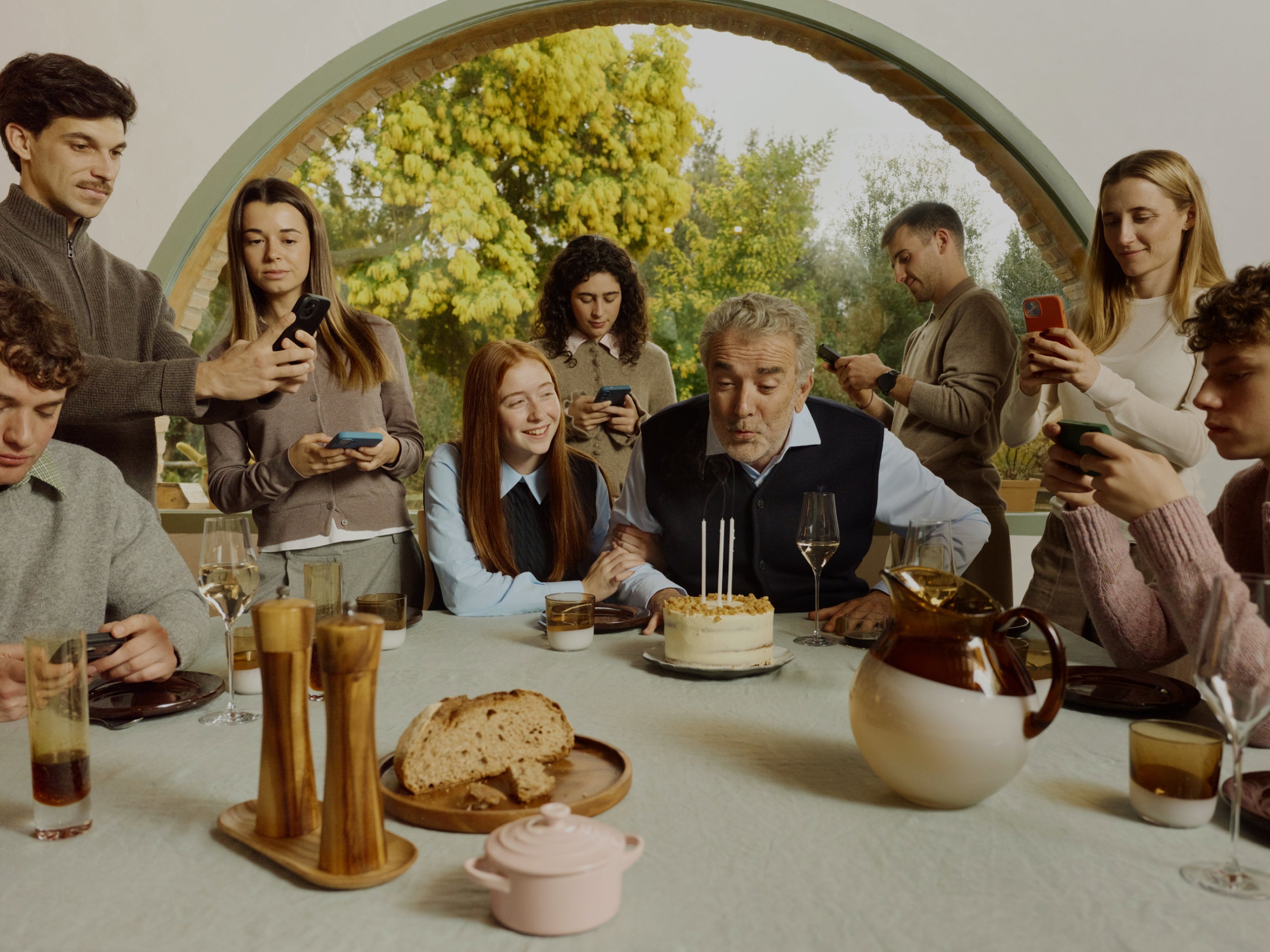 An older man blows out the candles on his birthday cake at a dining table while several people around him are distracted by their smartphones instead of watching.