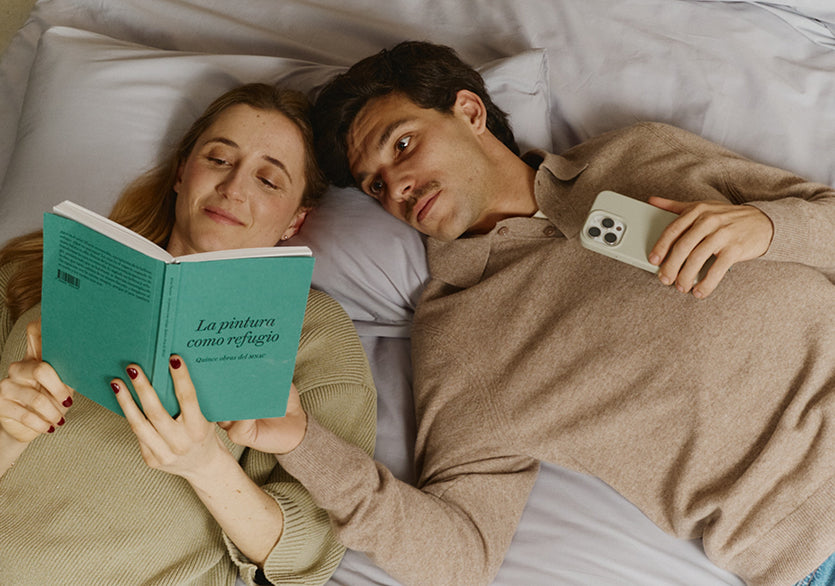 Couple lying in bed, woman reading a book, man using a phone