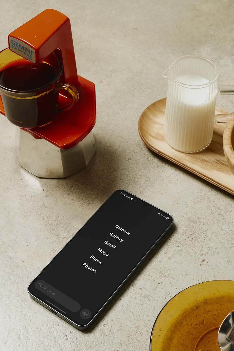 Smartphone on a light surface with a coffee maker, cup, and glass of milk in the background.